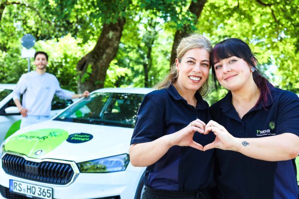 Team von 1gradmehr Pflege posiert mit den Firmenautos und bilden ein Herz mit Händen zwei Mitarbeiterinnen.